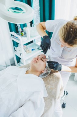 Klinikteki bir cildiyeci ya da kozmetikçi kadın hastalarla çalışır ve genç bir kadının yüzünde yüz bakımı yapar. Kozmetik uzmanının ofisindeki yüz ultrasonik temizleme.