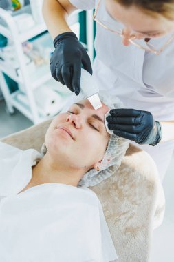 Klinikteki bir cildiyeci ya da kozmetikçi kadın hastalarla çalışır ve genç bir kadının yüzünde yüz bakımı yapar. Kozmetik uzmanının ofisindeki yüz ultrasonik temizleme.