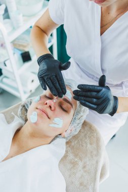 Bir güzellik uzmanı bir kadının yüzüne maske takar. Yüz bakımı. Klinikte bir cildiyeci ya da kozmetikçi kadın hastalarla çalışır ve genç bir kadının yüzünde yüz bakımı yapar..