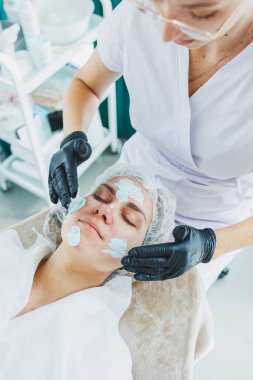 Bir güzellik uzmanı bir kadının yüzüne maske takar. Yüz bakımı. Klinikte bir cildiyeci ya da kozmetikçi kadın hastalarla çalışır ve genç bir kadının yüzünde yüz bakımı yapar..