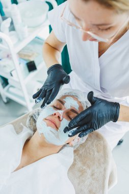 Bir güzellik uzmanı bir kadının yüzüne maske takar. Yüz bakımı. Klinikte bir cildiyeci ya da kozmetikçi kadın hastalarla çalışır ve genç bir kadının yüzünde yüz bakımı yapar..