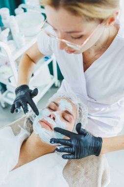 Bir güzellik uzmanı bir kadının yüzüne maske takar. Yüz bakımı. Klinikte bir cildiyeci ya da kozmetikçi kadın hastalarla çalışır ve genç bir kadının yüzünde yüz bakımı yapar..