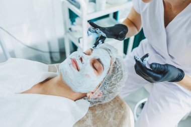 Güzellik uzmanının ofisi bir kaplıca salonundaki bir kadının yüzüne maske takıyor, yukarısı manzaralı. Estetik prosedür. Yüz derisi için bakım maskesi.