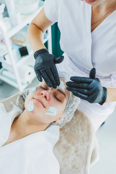 Bir güzellik uzmanı bir kadının yüzüne maske takar. Yüz bakımı. Klinikte bir cildiyeci ya da kozmetikçi kadın hastalarla çalışır ve genç bir kadının yüzünde yüz bakımı yapar..