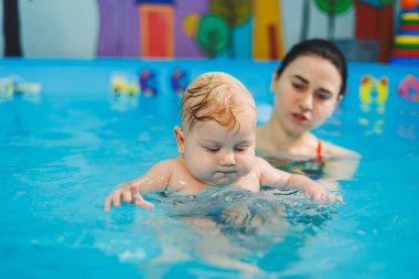 Bir bayan antrenör bebeğe havuzda yüzmeyi öğretir ve ellerini destekler. Havuzda yüzen bir bebek. Yeni doğmuş bir çocuğa havuzda yüzmeyi öğretiyorum.