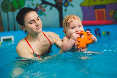 Bir bayan antrenör bebeğe havuzda yüzmeyi öğretir ve ellerini destekler. Havuzda yüzen bir bebek. Yeni doğmuş bir çocuğa havuzda yüzmeyi öğretiyorum.