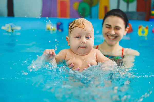 A female trainer teaches a baby to swim in the pool and supports his hands. Baby swimming in the pool. Teaching a newborn boy to swim in the pool