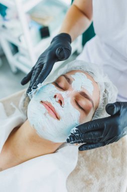 Bir güzellik uzmanı bir kadının yüzüne maske takar. Yüz bakımı. Klinikte bir cildiyeci ya da kozmetikçi kadın hastalarla çalışır ve genç bir kadının yüzünde yüz bakımı yapar..