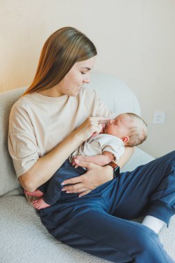 Gülümseyen genç bir anne bir kanepede oturuyor ve kucağında yeni doğmuş bir bebek var. Kollarında çocuğu olan mutlu bir anne. Annelik kavramı.