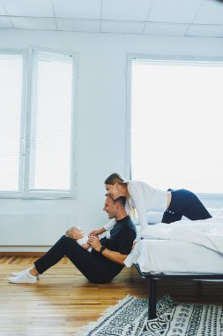 Evde mutlu bir aile: baba, anne ve yeni doğan oğul yatak odasındaki beyaz yatakta oynuyorlar. Genç aile birlikte eğleniyor, birbirlerine sarılıyorlar..