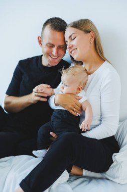 Genç anne ve baba, yeni doğan oğullarıyla beyaz bir yatakta oynuyorlar. Küçük çocuklu şık bir aile.