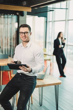 Beyaz gömlekli genç bir adam geniş, modern bir ofiste kahve içiyor ve tablet üzerinde çalışıyor. Modern ofis çalışanı. Bardaklı iş adamı internette çalışıyor ve rahatça kahve içiyor.