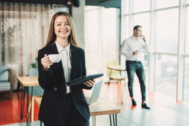 Siyah klasik takım elbiseli genç bir kadın modern bir ofiste dikilirken kahve içiyor. Ofiste portatif bir tablet üzerinde çalış..