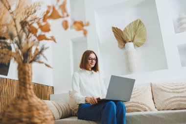 Genç serbest çalışan kadın kanepede oturuyor ve dizüstü bilgisayarda çalışıyor, dizüstü bilgisayarda film izliyor, çalışıyor, blog yazıyor, rahatlıyor ve internette sohbet ediyor..