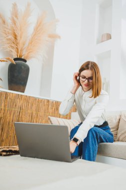 Gündelik giysiler içinde sevimli bir genç kadın evdeki kanepede oturuyor ve bir dizüstü bilgisayar üzerinde çalışıyor. Çevrimiçi olarak çalış.