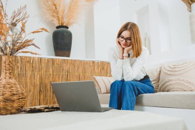 Genç serbest çalışan kadın kanepede oturuyor ve dizüstü bilgisayarda çalışıyor, dizüstü bilgisayarda film izliyor, çalışıyor, blog yazıyor, rahatlıyor ve internette sohbet ediyor..