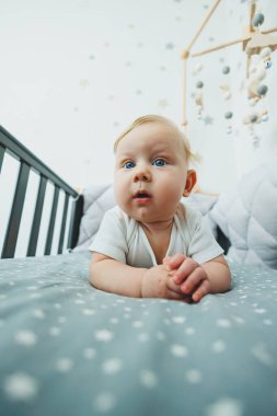 Beşikte yatan 5 aylık bir bebeğin fotoğrafı var. Beyaz elbiseli küçük bir çocukla bir karyola..