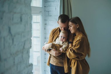 Mutlu bir anne ve baba, 4 aylık oğullarını kollarına alıp evlerinin penceresinden dışarı bakarlar. Yeni doğmuş bir bebekle mutlu ebeveynlerin ev yapımı fotoğrafı. Aile kavramı