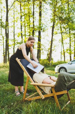 Bir aile çifti, bir erkek ve bir kadın, doğada dinleniyorlar ve bir dizüstü bilgisayarla uzaktan çalışıyorlar. İnternette çalışan sevgili çiftlerin pikniği. Hamile bir kadın kocasıyla birlikte doğada dinleniyor.