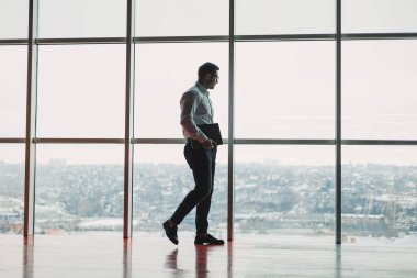 Dizüstü bilgisayarı olan genç yakışıklı bir adam gökyüzündeki büyük bir panoramik pencerenin arka planında duruyor. Geniş pencereli ve ofis çalışanı olan modern bir ofis.