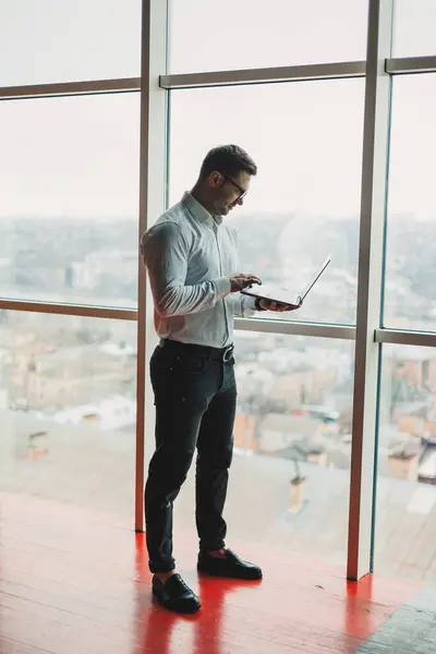 A young handsome man with a laptop stands on the background of a large panoramic window in the sky. A modern office with large windows and an office worker
