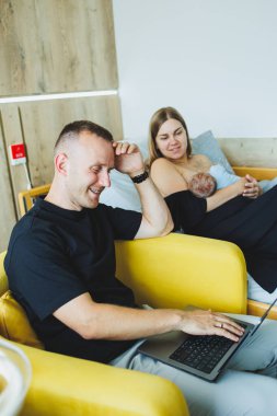Uzaktan kumandalı internet. Karısı çocuğa bakarken mutlu bir baba dizüstü bilgisayarda çalışıyor. Evden çalışmak, ebeveynlik ve uzaktan kumanda. Yeni doğmuş bir bebeği emziriyor.