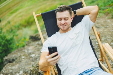 Sakallı bir adam doğada katlanır ahşap bir sandalyede oturur ve telefonda konuşur. Sadece doğadaki eğlence kavramı..