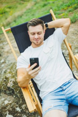 Sakallı bir adam doğada katlanır ahşap bir sandalyede oturur ve telefonda konuşur. Sadece doğadaki eğlence kavramı..