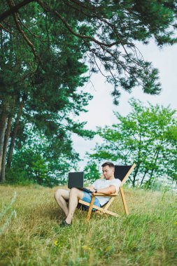 Katlanan ahşap bir sandalyede oturan bir adam doğada oturur ve uzaktan bilgisayarla çalışır. Sadece doğadaki eğlence kavramı..