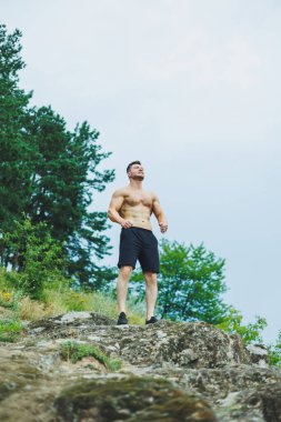 Genç kaslı bir adam ormanın yakınındaki kayalıklarda oturuyor. Rahatlatıcı ve kaslı bir erkek vücudu..