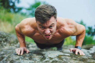 Genç kaslı bir adam ormanın yakınındaki kayalıklarda oturuyor. Rahatlatıcı ve kaslı bir erkek vücudu..