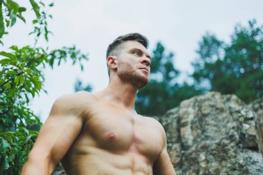 Genç kaslı bir adam ormanın yakınındaki kayalıklarda oturuyor. Rahatlatıcı ve kaslı bir erkek vücudu..