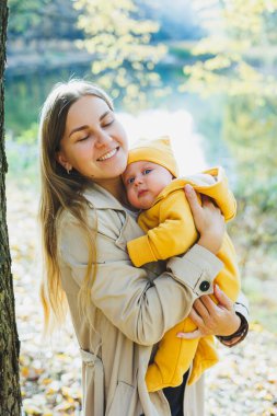 Güneşli bir parkta dururken küçük oğlunu kollarında tutan gülümseyen bir anne. Doğada yeni doğmuş bir bebekle yürüyüş..