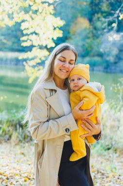 Güneşli bir parkta dururken küçük oğlunu kollarında tutan gülümseyen bir anne. Doğada yeni doğmuş bir bebekle yürüyüş..
