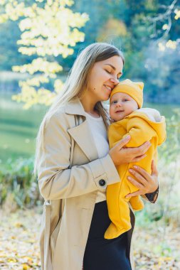 Güneşli bir parkta dururken küçük oğlunu kollarında tutan gülümseyen bir anne. Doğada yeni doğmuş bir bebekle yürüyüş..
