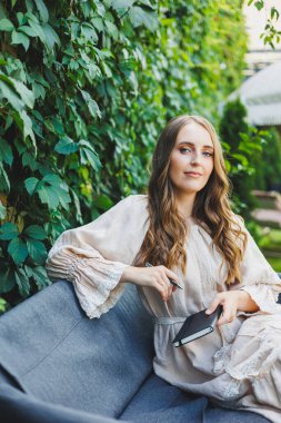 Elbiseli hoş bir genç kadın elinde bir defterle oturur ve bir yaz kafesinin terasına notlar alır. Gündelik şık yaz kıyafetleri içinde bir kadın.