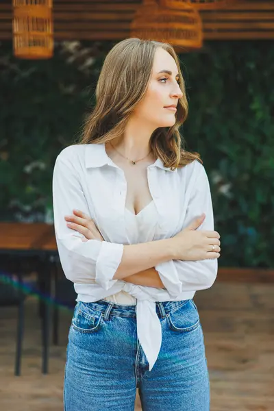 A cute young woman in a white shirt and blue jeans is standing on the terrace of a summer cafe. Woman in casual stylish summer clothes