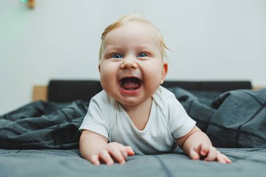 Beyaz elbiseli dört aylık küçük bir çocuk yatakta uzanıyor ve gülümsüyor. Gülümseyen bir bebek