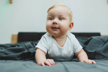 Beyaz elbiseli dört aylık küçük bir çocuk yatakta uzanıyor ve gülümsüyor. Gülümseyen bir bebek