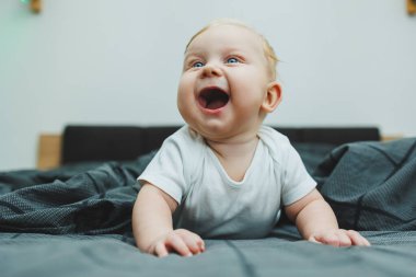 Beyaz elbiseli dört aylık küçük bir çocuk yatakta uzanıyor ve gülümsüyor. Gülümseyen bir bebek