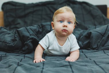 Beyaz elbiseli dört aylık küçük bir çocuk yatakta uzanıyor ve gülümsüyor. Gülümseyen bir bebek