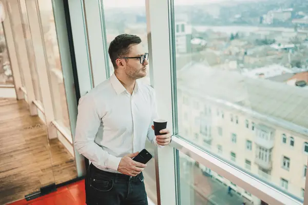 A male manager in a white shirt and glasses stands in the office by the window and drinks coffee while watching a video on the phone. Business telephone conversation of a business man.