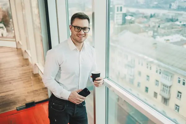 A male manager in a white shirt and glasses stands in the office by the window and drinks coffee while watching a video on the phone. Business telephone conversation of a business man.