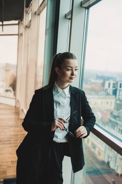 Siyah takım elbiseli, gözlüklü, modern bir iş kadını pencerenin yanında duruyor ve metropol 'e bakıyor. Gözlüklü ve beyaz gömlekli kadın yönetici..