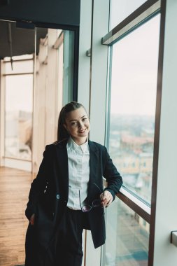 Siyah takım elbiseli, gözlüklü, modern bir iş kadını pencerenin yanında duruyor ve metropol 'e bakıyor. Gözlüklü ve beyaz gömlekli kadın yönetici..