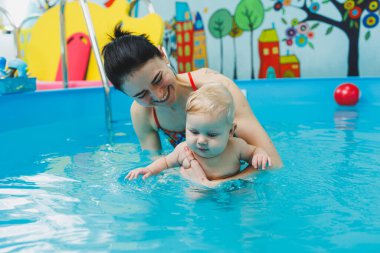 6 aylık bir bebek havuzda koçla yüzmeyi öğreniyor. Bebekler için yüzme havuzu. Çocuk gelişimi