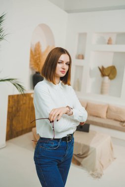 Gözlüklü güzel bir kadın parlak bir salonun ortasında duruyor, beyaz bir tişört ve kot pantolon giyiyor. Uzaktan çalışan kendine güvenen genç bir kadın..