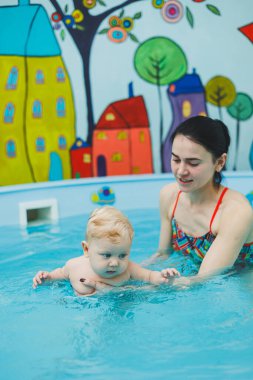 6 aylık bir çocuk havuzda yüzüyor. Antrenör bebeğe yüzme, çocuklar için yüzme havuzu öğretiyor. Çocuk gelişimi.