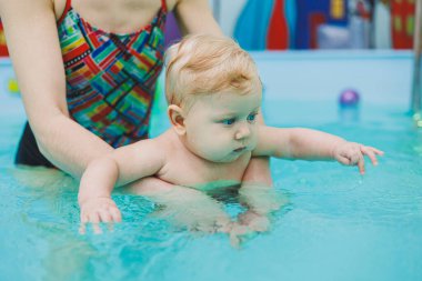 6 aylık bir çocuk havuzda yüzüyor. Antrenör bebeğe yüzme, çocuklar için yüzme havuzu öğretiyor. Çocuk gelişimi.