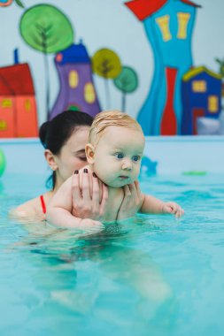 6 aylık bir çocuk havuzda yüzüyor. Antrenör bebeğe yüzme, çocuklar için yüzme havuzu öğretiyor. Çocuk gelişimi.